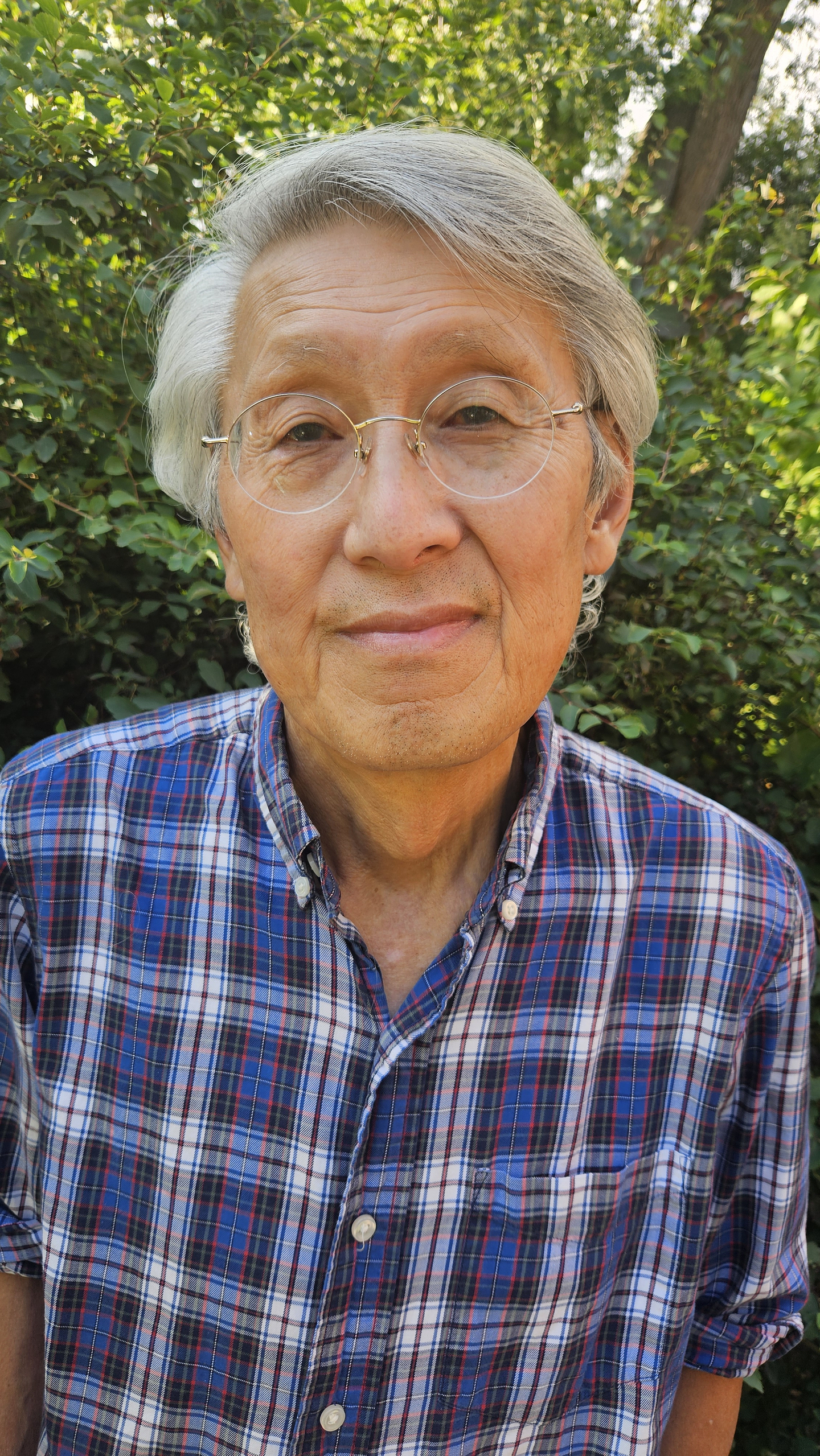 Standing against lush foliage, Gerry Shikatani gazes at the camera with a closed-mouth smile. He wears round rimless glasses and a blue and red plaid shirt. 