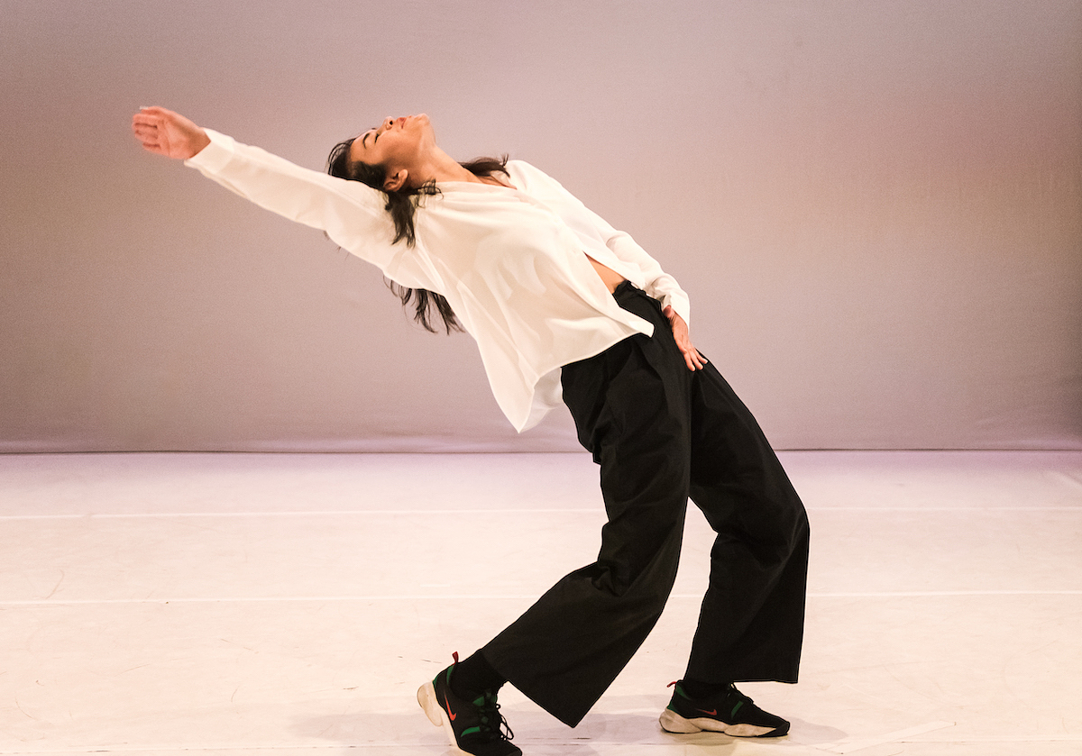 Amrita Hepi dances on a white stage brightly illuminated by soft pink lighting. Wearing a white shirt, black trousers, and sneakers, her torso is tilted backwards so that her face is upturned towards the sky. Her right arm is outstretched behind her and her left hand rests on her hip.