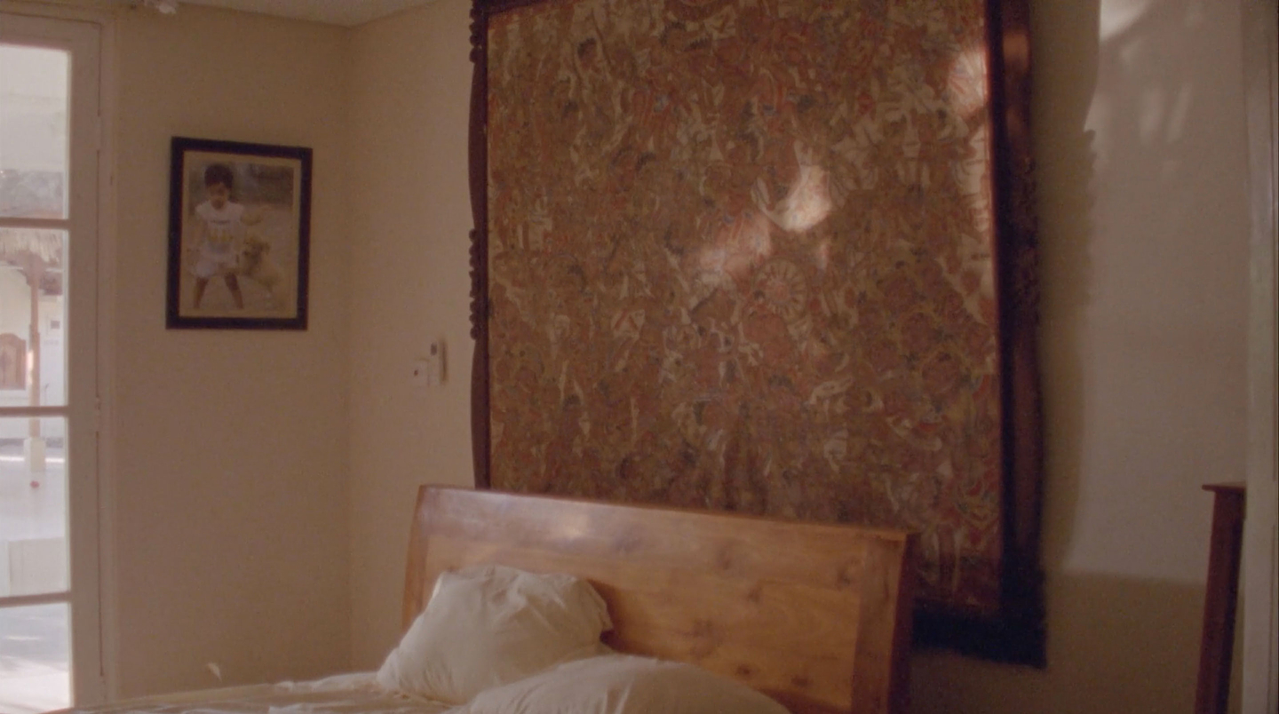 An interior view of the corner of a bedroom. A bed’s wooden headboard and two white pillows appear in front of a large patterned fabric painting in a wooden frame on the wall. A small framed photograph of a young boy with a dog hangs on the perpendicular wall, next to a French door.