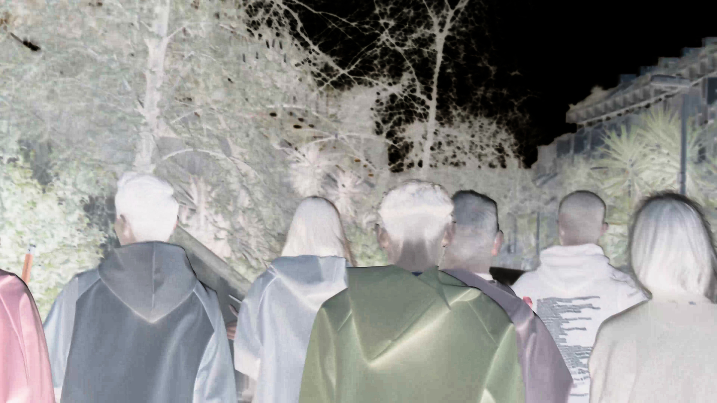 The image shows a group of people walking outdoors at night, viewed from behind, with the colors inverted so it appears like a photographic negative. The figures wear jackets or hooded garments, and they move along a path bordered by dense trees on the left and a building with plants or palm trees on the right.

