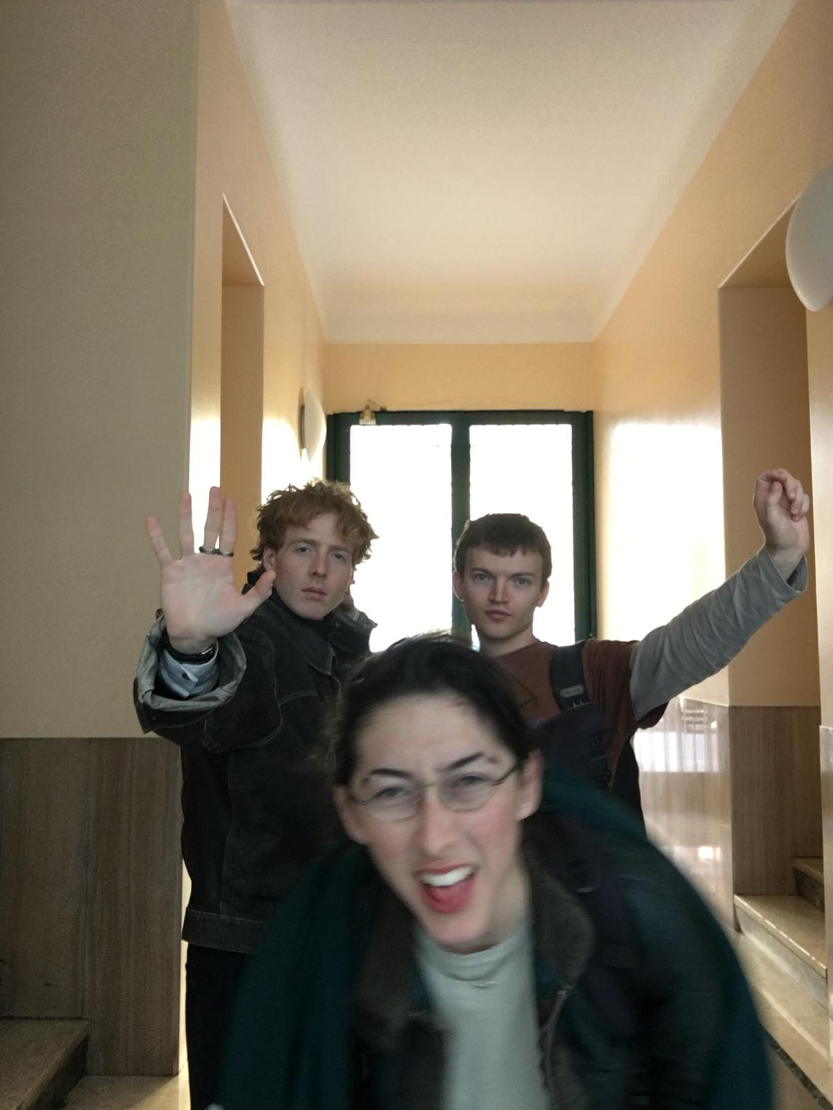 Finlay Clark and David Kenny stand with their hands raised behind Jess Hickie-Kallenbach, who crouches in fron, smiling and blurry from motion. The bandmates are posed in a brightly lit hallway. 