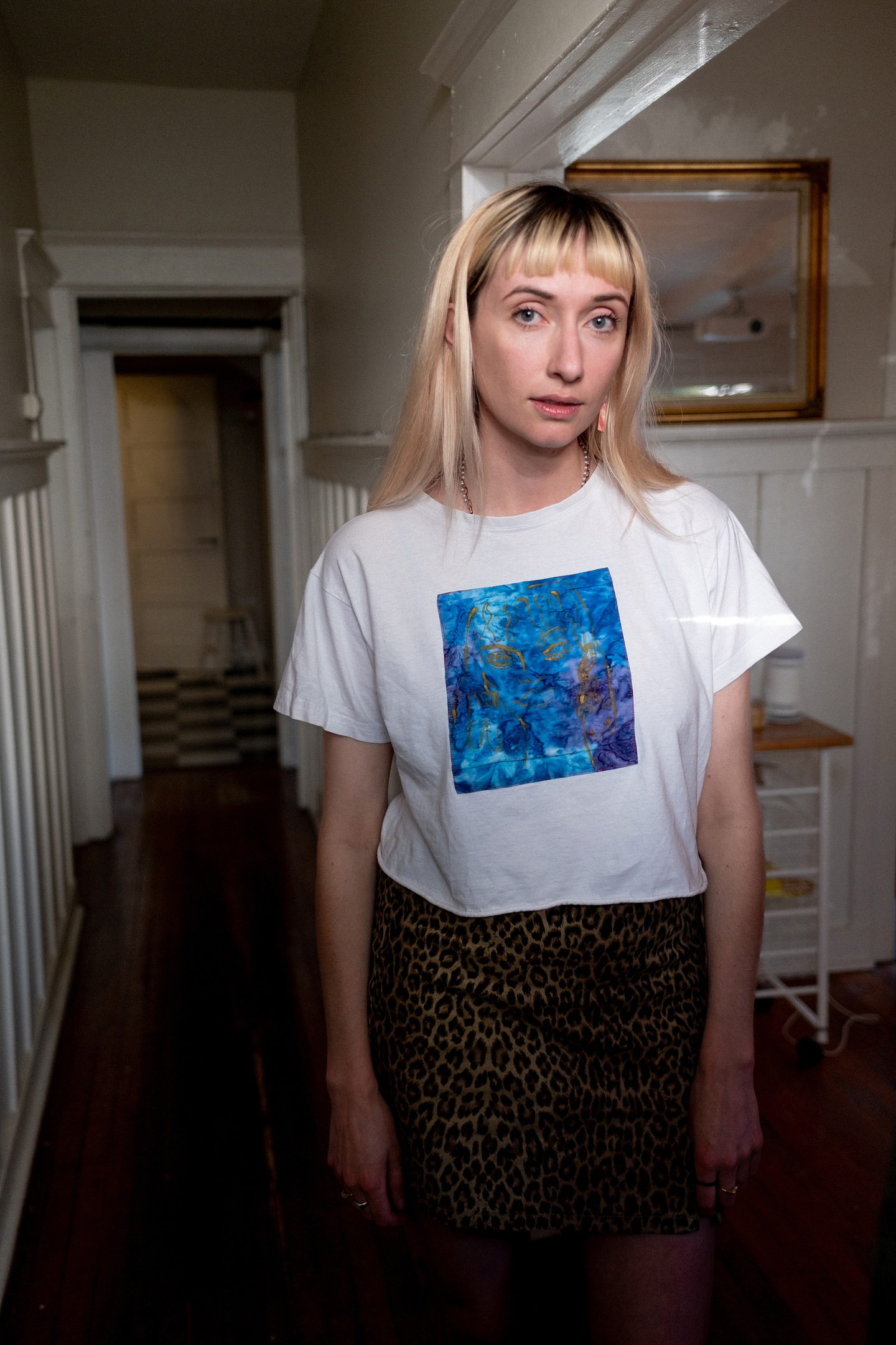 Darragh Amelia stands indoors in a hallway with wooden floors and white paneling. She is wearing a white t-shirt featuring a blue abstract artwork on the front and a leopard print skirt. A framed mirror hangs on the wall behind her, and a narrow utility cart is visible to the side. The lighting highlights her calm, direct expression.