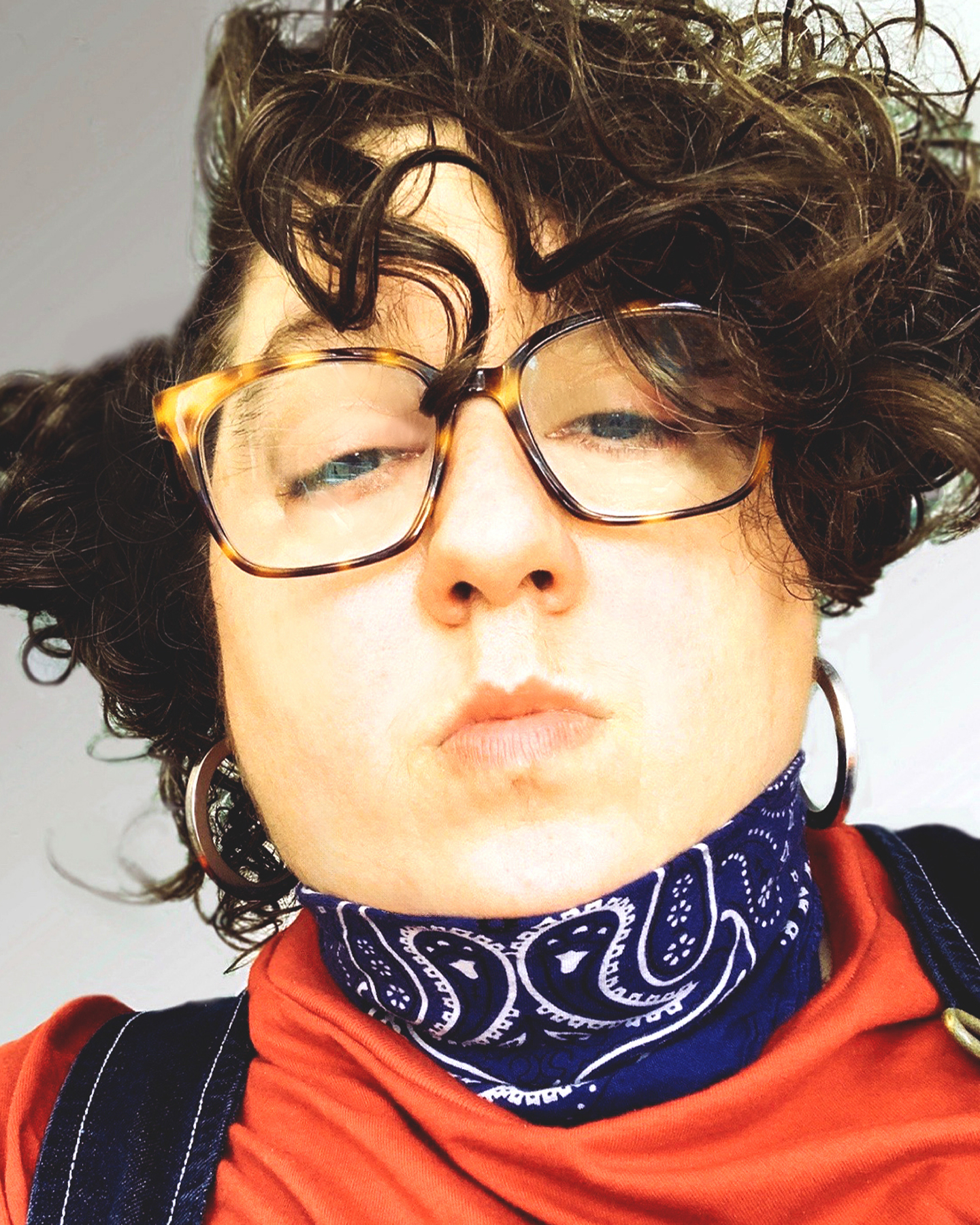 Close-up, low-angle selfie of Constantina Zavitsanos with tousled dark curly hair, large tortoiseshell glasses and silver hoop earrings, eyes half-closed and lips slightly pursed. They wear a navy paisley bandana knotted at the neck, an orange-red T-shirt and dark denim overalls, against a pale wall.