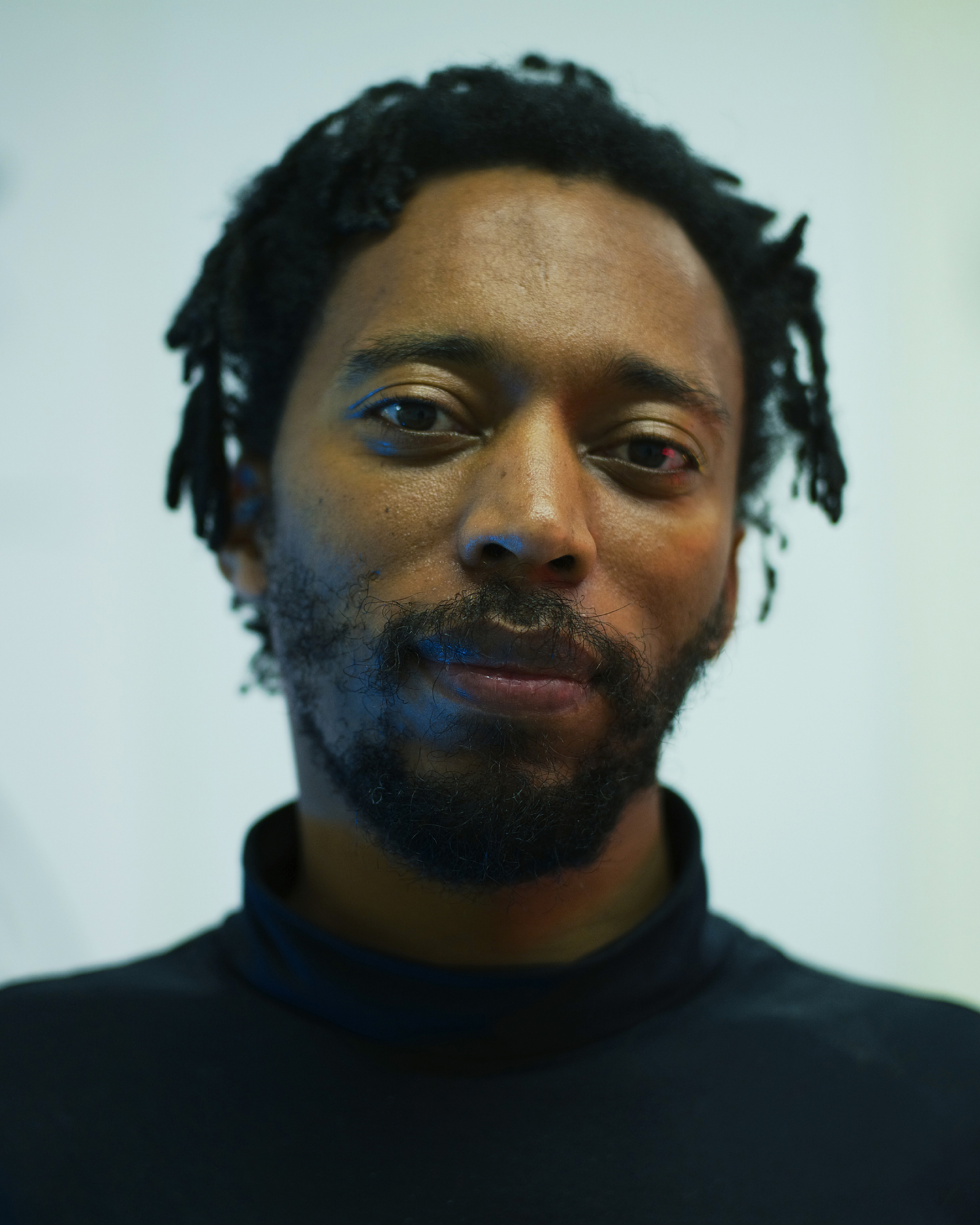 Portrait of Christian Nyampeta with short locs and a beard, wearing a black turtleneck and looking directly at the camera with a slight smile. Soft blue-green light falls across his face, and behind him, out of focus, are loose charcoal or ink drawings on a pale wall.