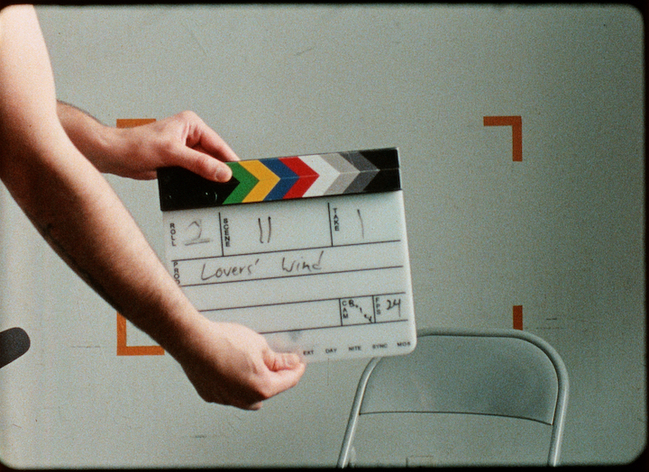 A clapperboard is held centre frame, featuring the words "Roll 2, Scene 11, Take 1, Lovers' Wind" in black texta.