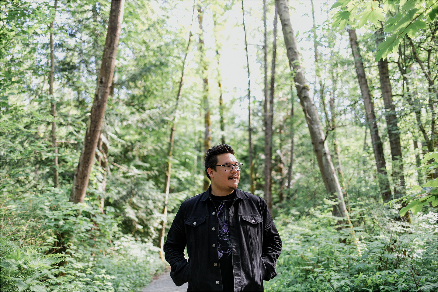 Gazing to the right, Jordan Abel stands on a path in a forest with his hands in the pockets of his black denim jacket. 