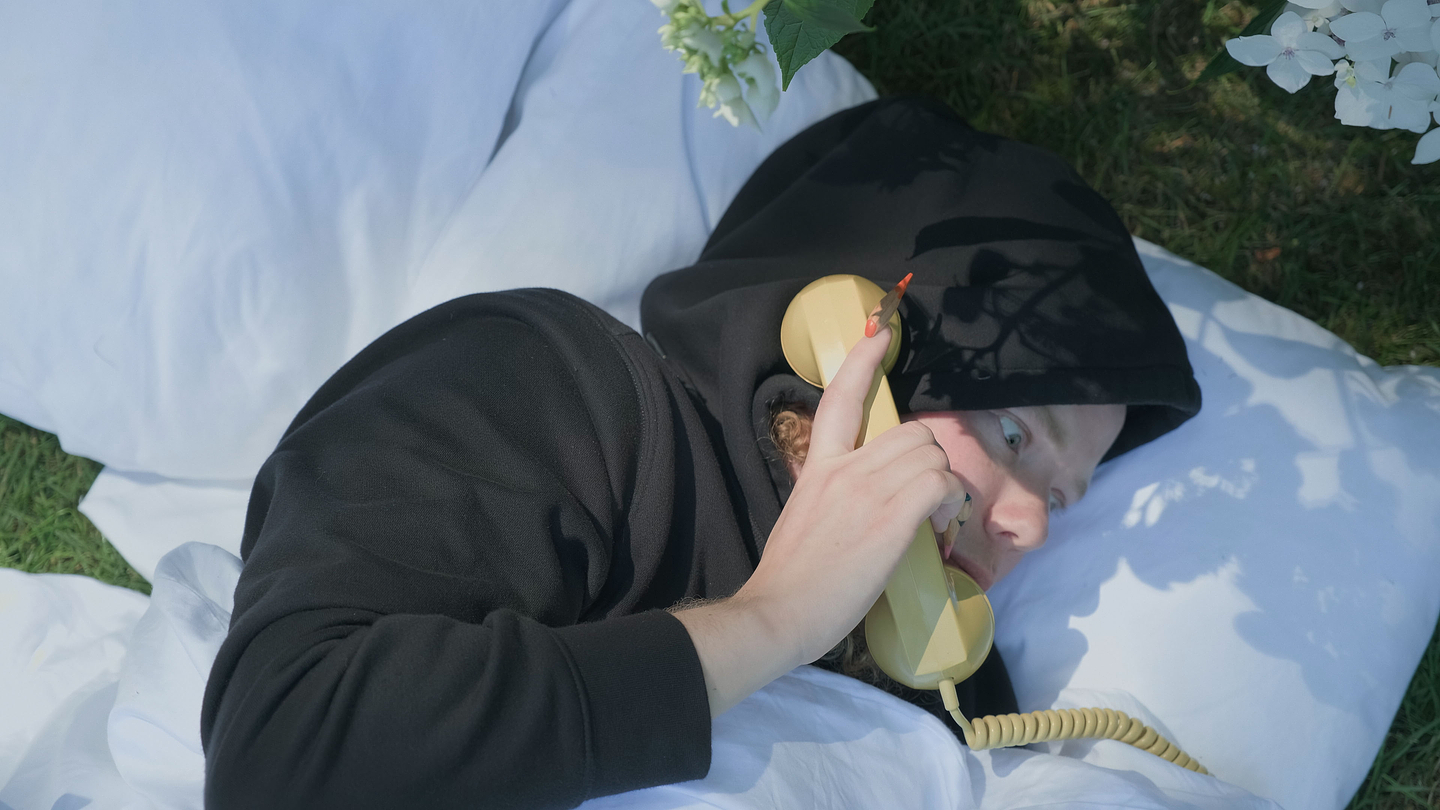 Archie Barry lying on white pillows outdoors, wearing a black hoodie, holding a yellow rotary phone receiver to their ear, with sunlight and flower shadows cast across their face.
