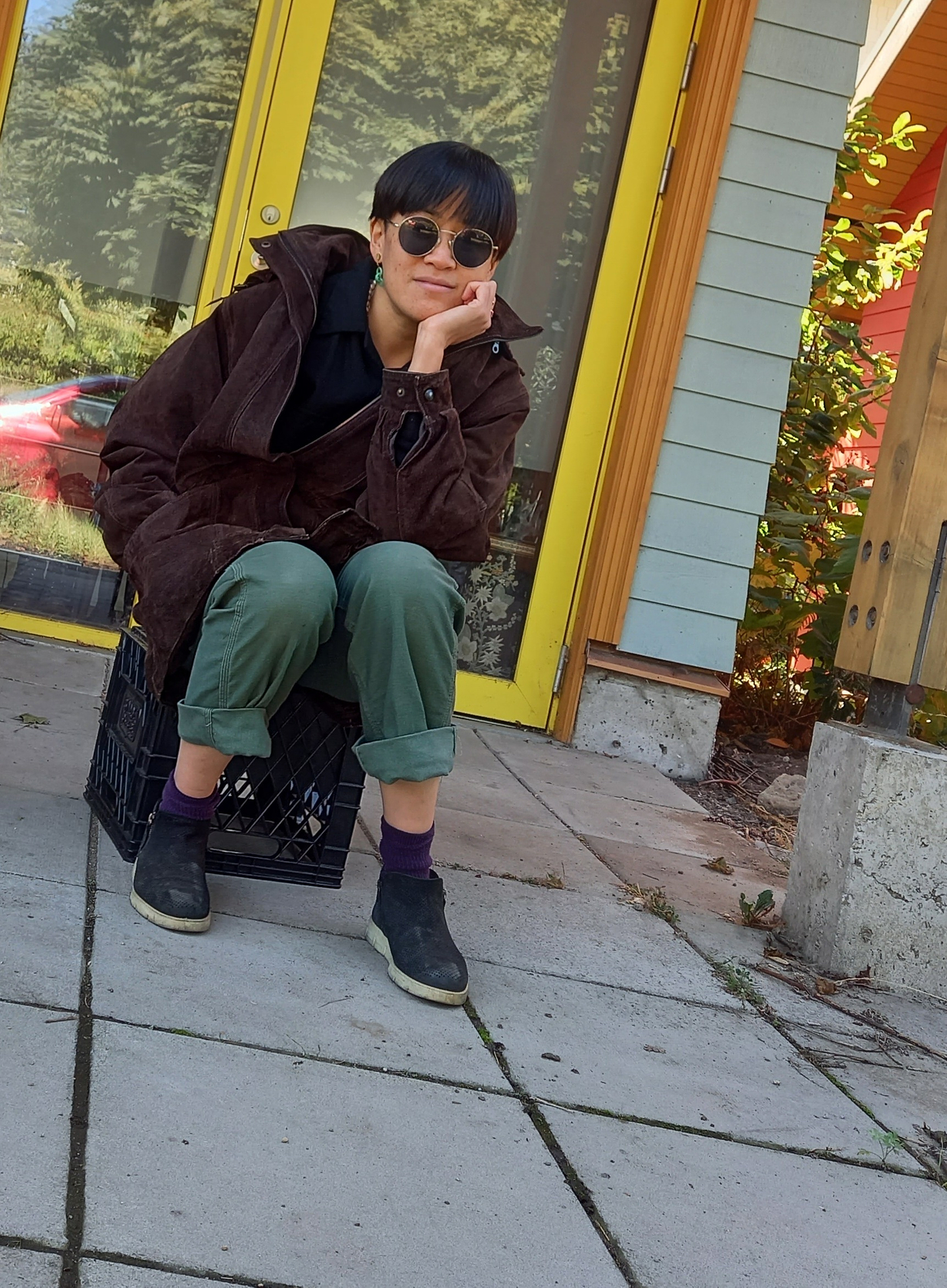 S F Ho sits on a black milk crate outside of a building with a glass door and yellow trim. They cradle their chin in their palm, and wear round sunglasses, a brown jacket, green cuffed pants, purple socks, and ankle boots.