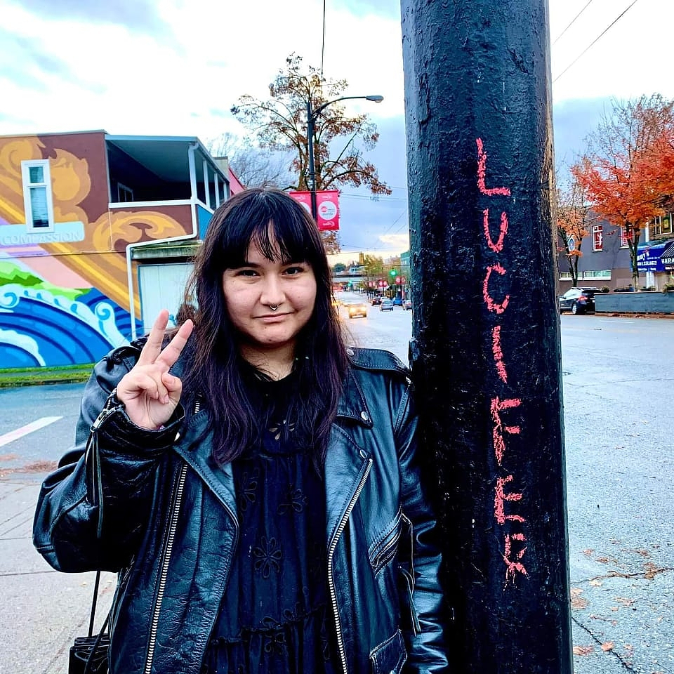 A portrait of Brandi Bird standing beside a black post with the word “Lucifer” written vertically in bright pink lettering. They are giving the peace sign with their right hand. In the background are trees with autumnal leaves and buildings with murals painted on their exterior walls.