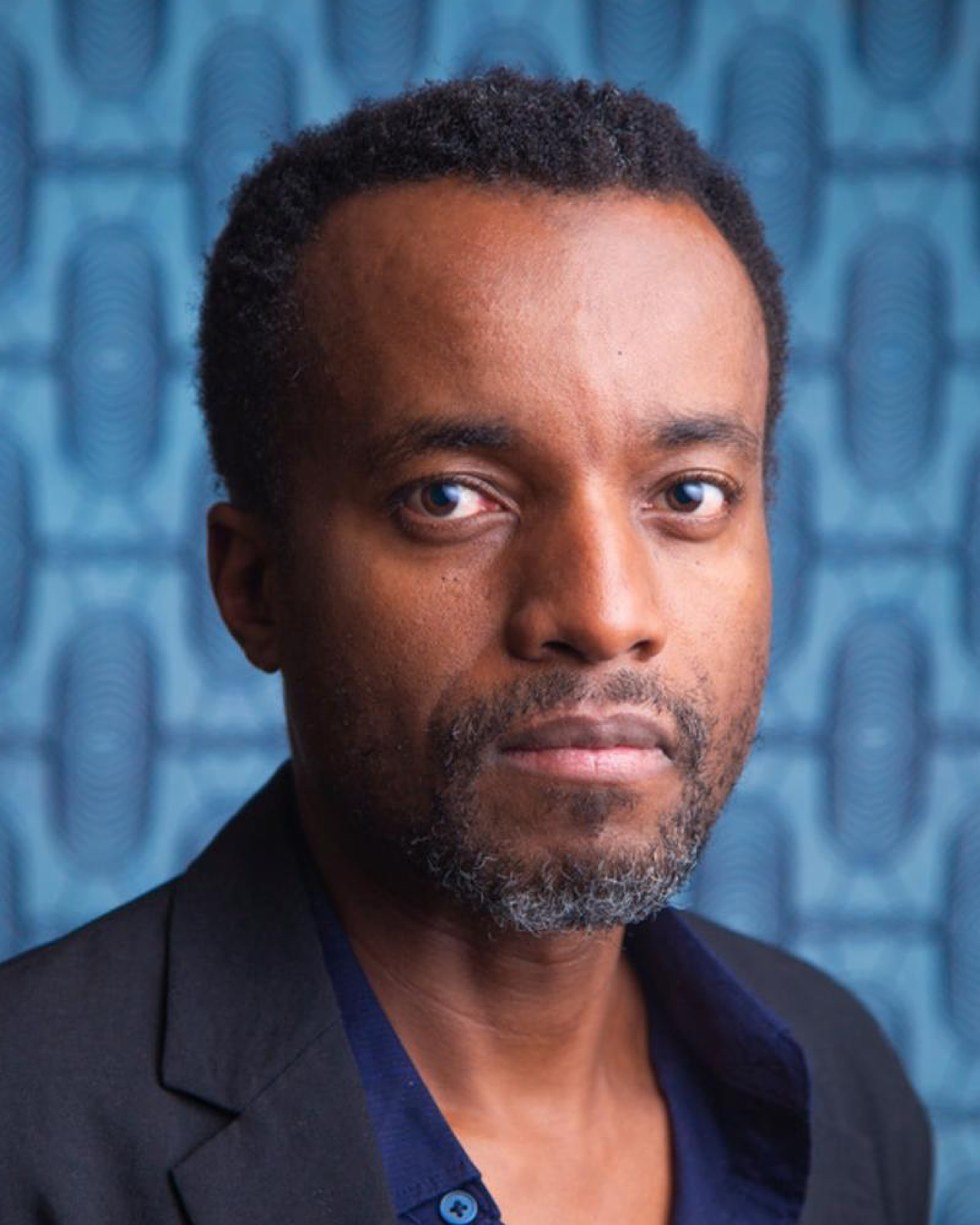 Portrait of Kivu Ruhorahoza with short dark hair and a greying beard, wearing a black blazer over an open-collared navy shirt and looking directly at the camera with a calm, steady expression. Behind him is a blue patterned backdrop of repeating oval, fingerprint-like motifs.