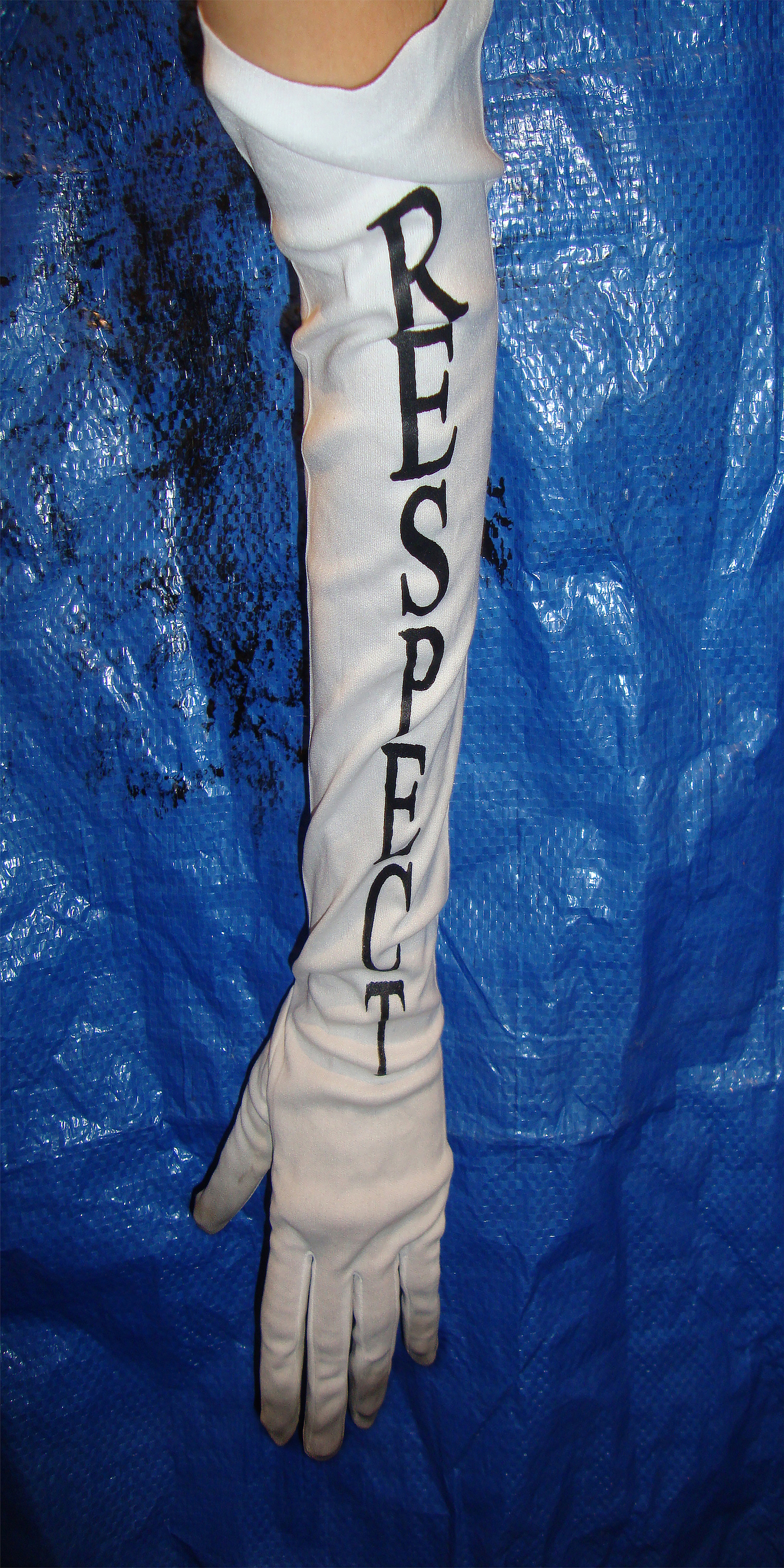 The word RESPECT is printed in black serif text on a white satin glove with dirty finger tips. The glove is worn by a disembodied arm stretched over a blue tarp background. 