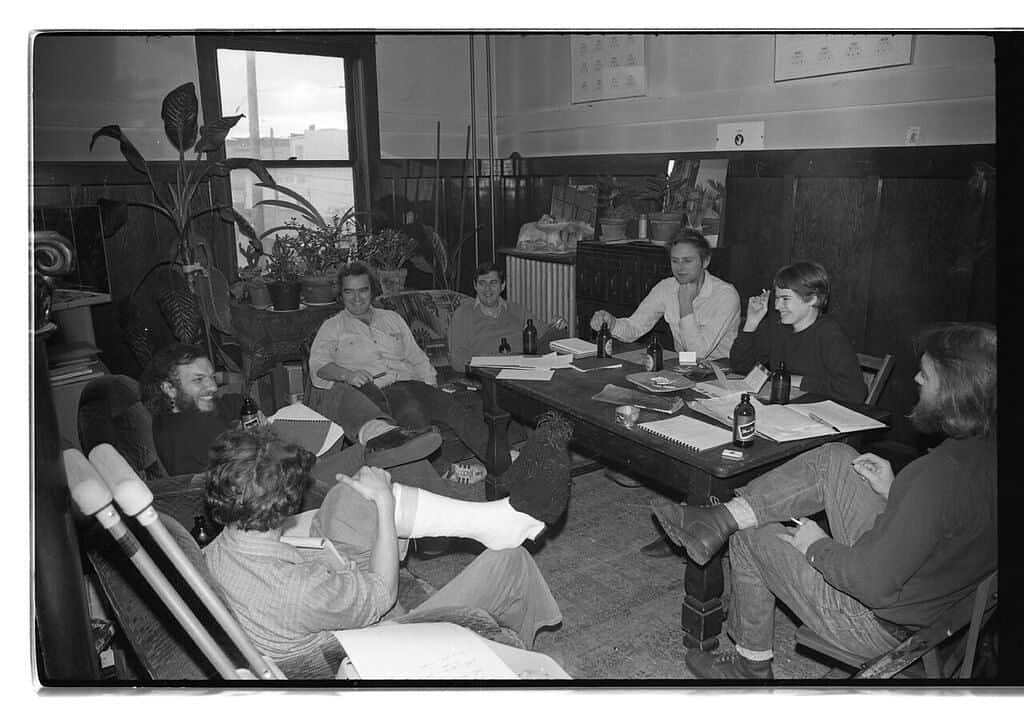 A black and white photograph of Western Front’s founders, relaxed and seated in a circle around a large wooden table in a room furnished with potted plants at Western Front.