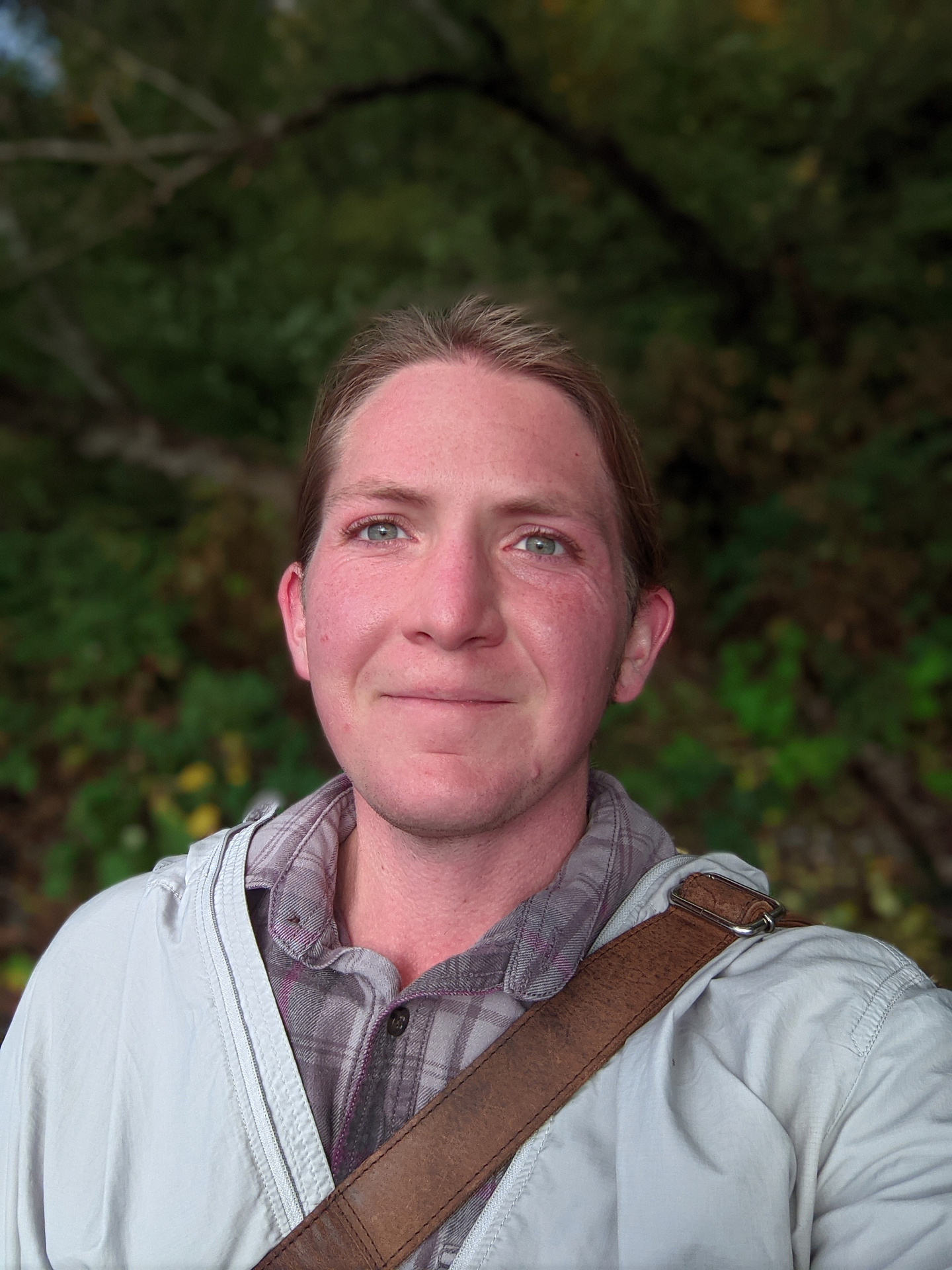 A selfie of Jay Pahre with a closed-mouth smile. His blonde hair is pulled back, and he wears a plaid shirt under a light grey windbreaker. A brown leather bag strap crosses his shoulder. Lush foliage can be seen in the background.