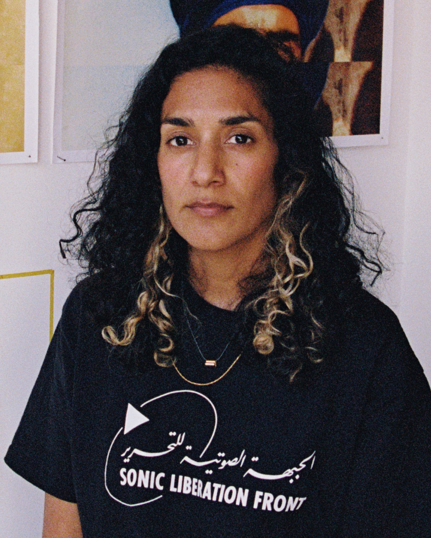 A portrait of Jasleen Kaur with long, dark curly hair with blonde highlights, seated on a stool in a white studio and looking directly at the camera with a calm expression. She wears a black T-shirt printed with "Sonic Liberation Front" in English and Arabic, gold necklaces and bracelets, and a patterned fabric wristband. Behind them hang two large prints, one repeating a close-up of heavily lined eyes in a vertical strip and the other repeating an image of a man in a blue turban. Small snapshots are taped inside yellow tape outlines on the wall, and a long green-and-pink knitted banner reading "LLOOOOOOON" hangs at the right.