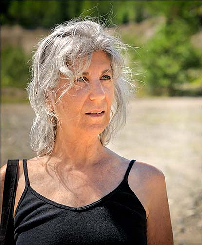 Rhoda Rosenfeld gazes beyond the camera with her head turned to her left. She has silver hair and wears a black spaghetti-strap tank top. A river and trees can be seen in the background.