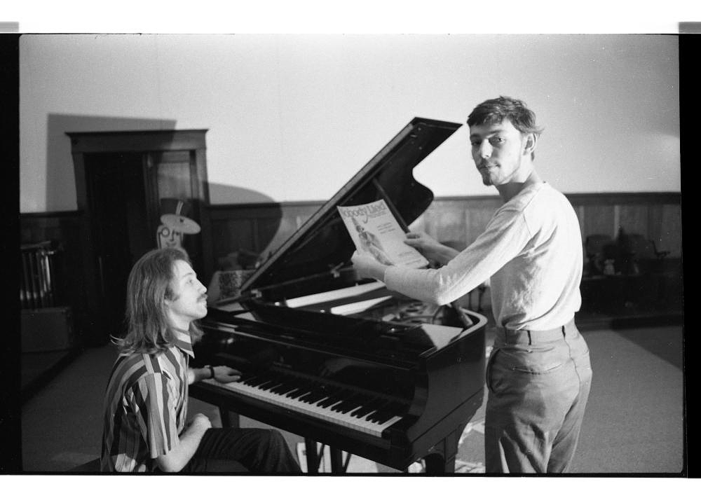 Hank Bull sits at a piano with his left hand on the keys. Pascal stands next to the piano holding sheet music with both hands and gazes back at the camera. The image is in black-and-white.
