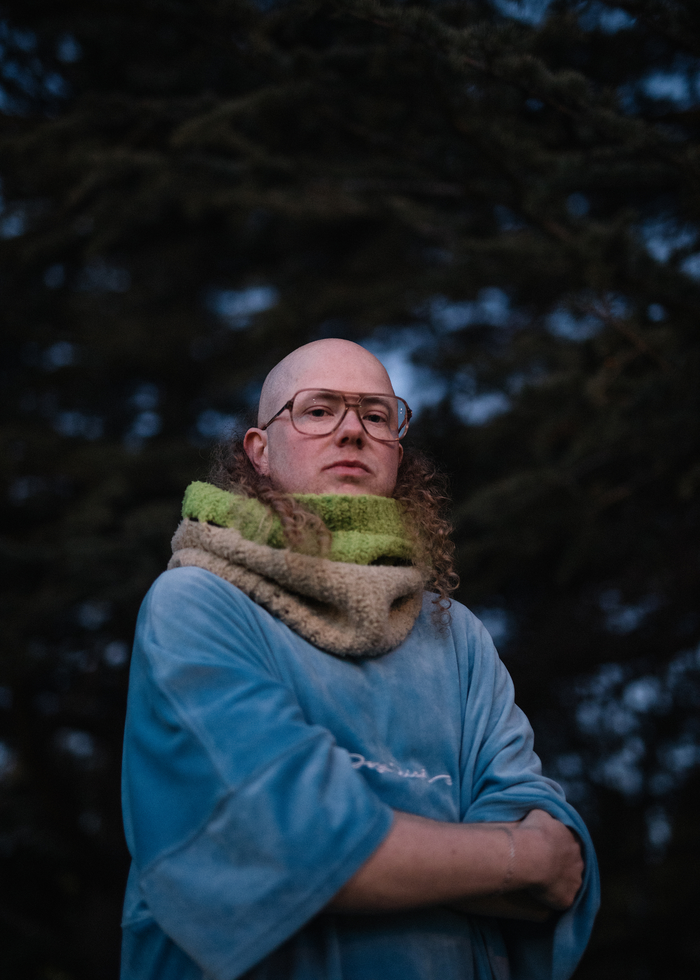 A person with a bald head and long curly side hair stands outdoors against a backdrop of dark trees. They wear tortoise shell aviator glasses, a loose blue velour top, and a chunky knitted scarf in beige and green. Their arms are folded, and they look directly at the camera with a calm, serious expression.
