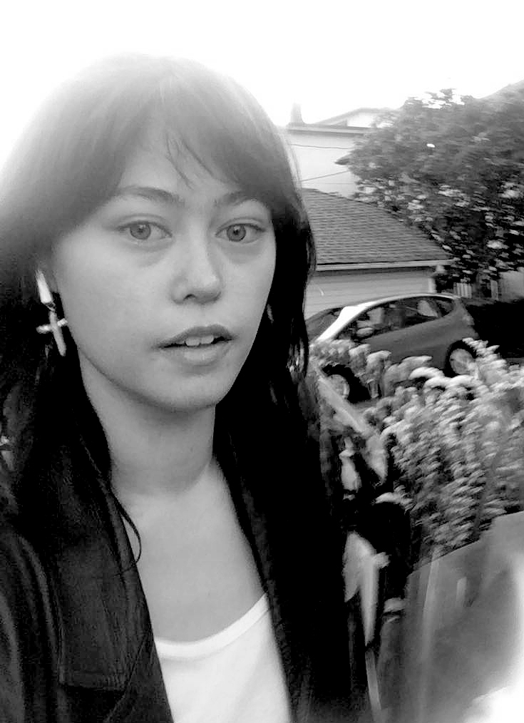 A portrait of Kiel Torres looking directly at the camera. She holds a bouquet of flowers and is standing in a suburban street, with a parked car and houses visible behind her. The brightness of the sun blows out the image at its edges.