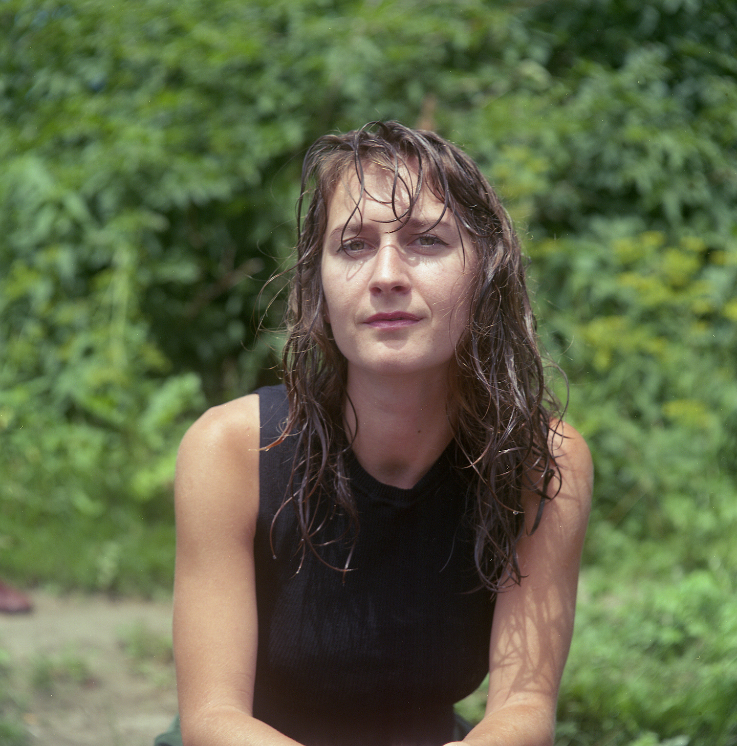 A photo of Ami Xherro with wet, shoulder-length dark hair sitting outdoors in front of blurred green foliage, wearing a black sleeveless top.