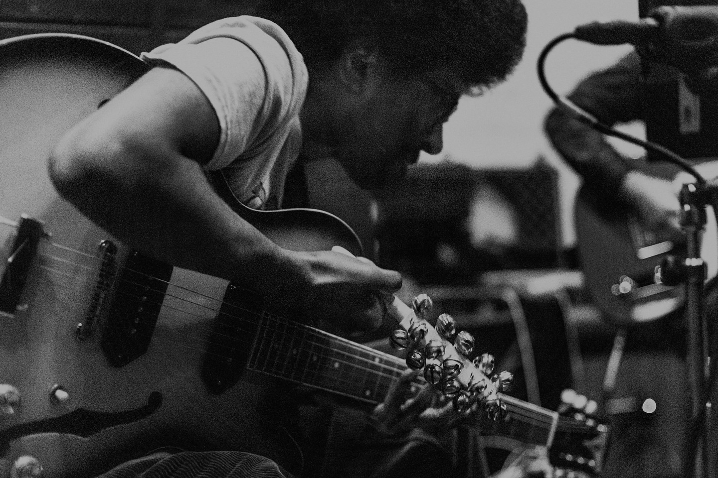 Seated, Mustafa Rafiq plays electric guitar while holding a jingle bell stick. The image is in black-and-white. 