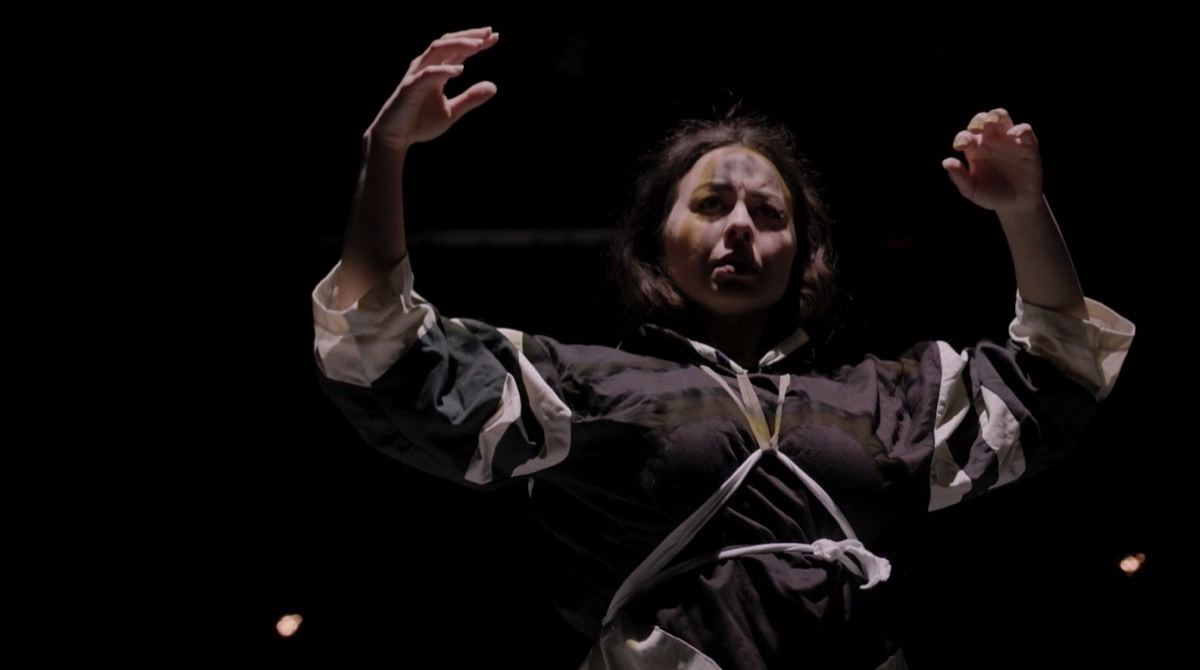 A photograph of Tiffany Ayalik performing Itqaqhaijuq / Tries to Remember at Western Front. Ayalik is wearing  an amaut – traditional Inuit woman’s coat – with black-and-white patterns, and a white rope-like belt tied across her chest and around her waist. Her arms form a U-shape around her face, and colourful, abstracted projections appear on her face.