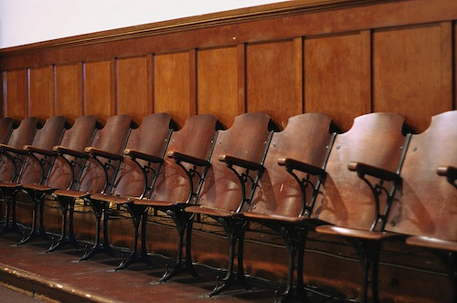 A row of built-in folding cottonwood chairs installed in front of wainscotting. All are painted in a rich reddish-brown stain.