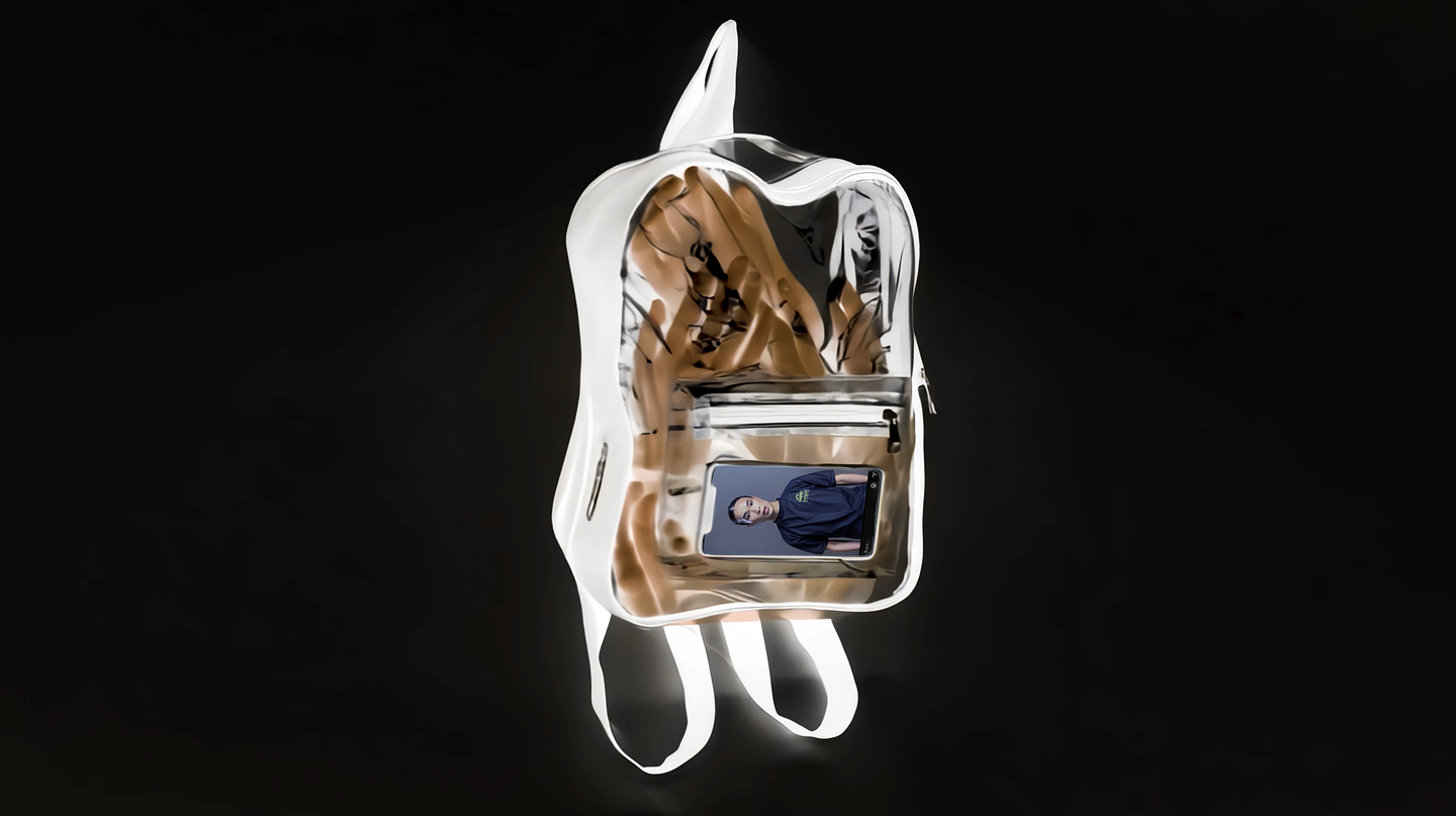 A clear backpack floating against a black background, with tan-colored hands or gloves visible inside and a smartphone in the front pocket displaying the portrait of a person in a dark shirt.