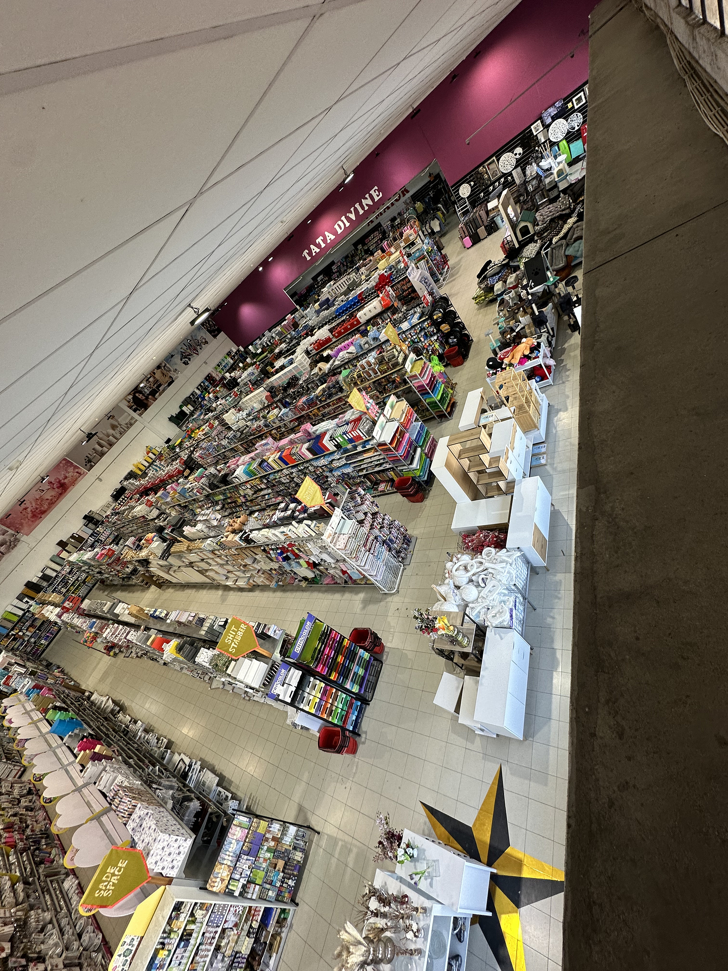 The aisles of a densely merchandised home goods store are seen from above. A large gold and black vinyl star is adhered to the beige tile floors beneath a display of cabinets. The far wall is painted burgundy with a sign reading Tata Divine in white serif text. 