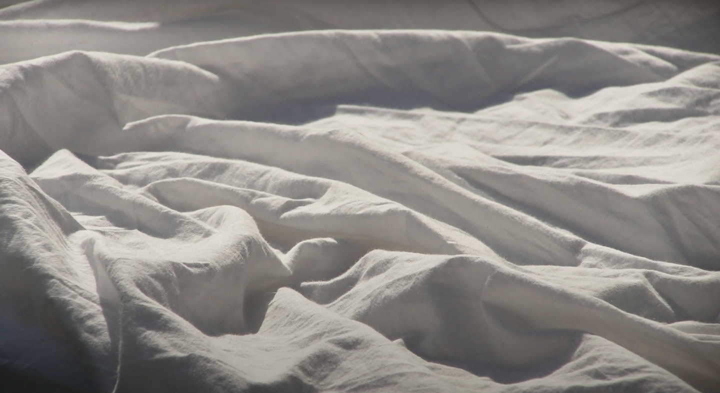 Soft, rumpled white bedsheets lit by gentle sunlight, forming wave-like folds and shadows across the surface.