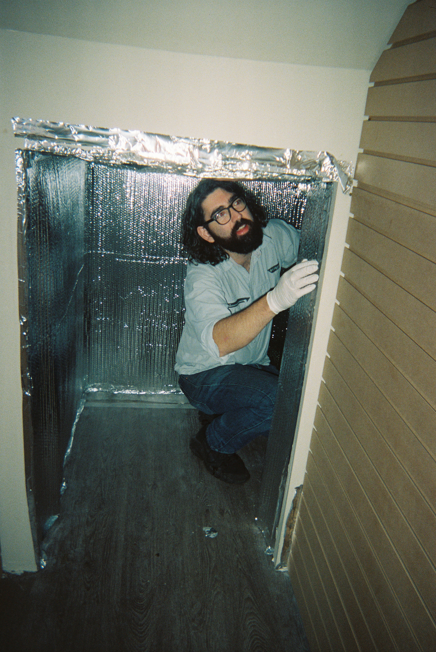 Sol Hashemi kneels in a small closet space with walls covered in silver-coloured reflective insulation. He wears a light blue shirt with dark blue jeans and a white glove on his hand.