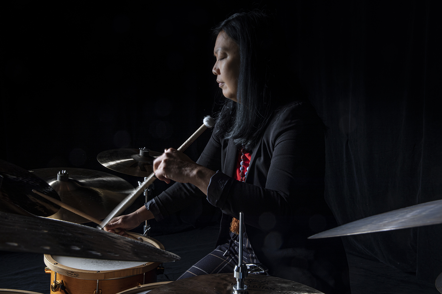 Susie Ibarra plays a drum set in a dimly lit room with a black backdrop. She is mid-performance, eyes closed, holding mallets and striking the cymbals and drum. She has long dark hair and wears a black blazer over a red top. The lighting emphasizes her focused expression and the reflective surfaces of the instruments.