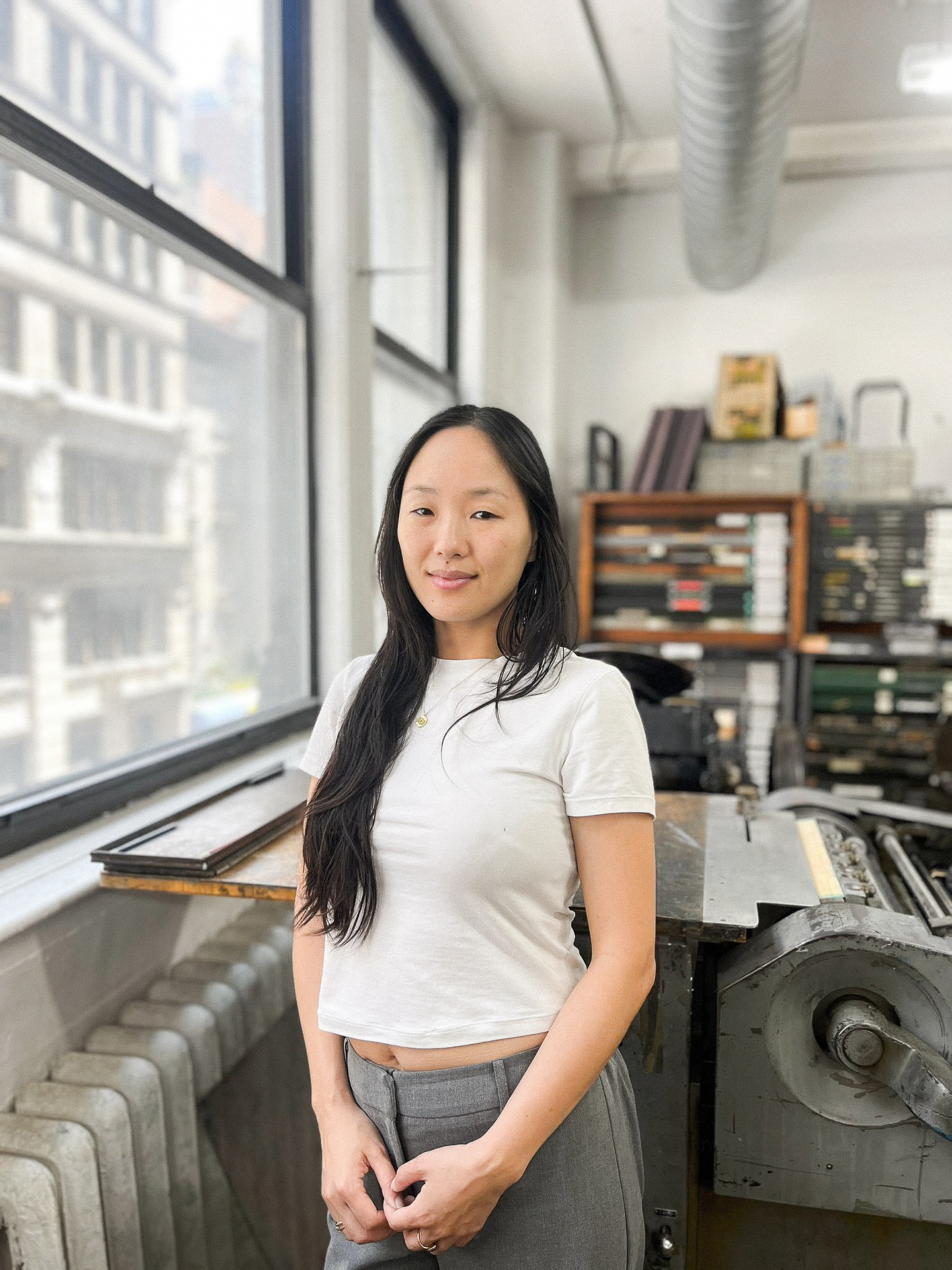 Rebecca Yeong Ae Mzengi Corey stands indoors near large industrial windows in a well-lit room filled with vintage printing equipment and metal type drawers. She is wearing a white cropped t-shirt and high-waisted gray pants, with a small gold necklace. She has a calm and confident expression, and natural light streams in through the windows, illuminating the room and cityscape outside.