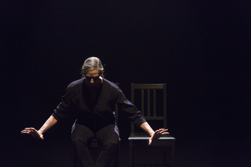 A colour photograph of Peggy Baker seated on stage. She looks down towards the floor with her arms outstretched to her sides. To her right is an empty black chair.