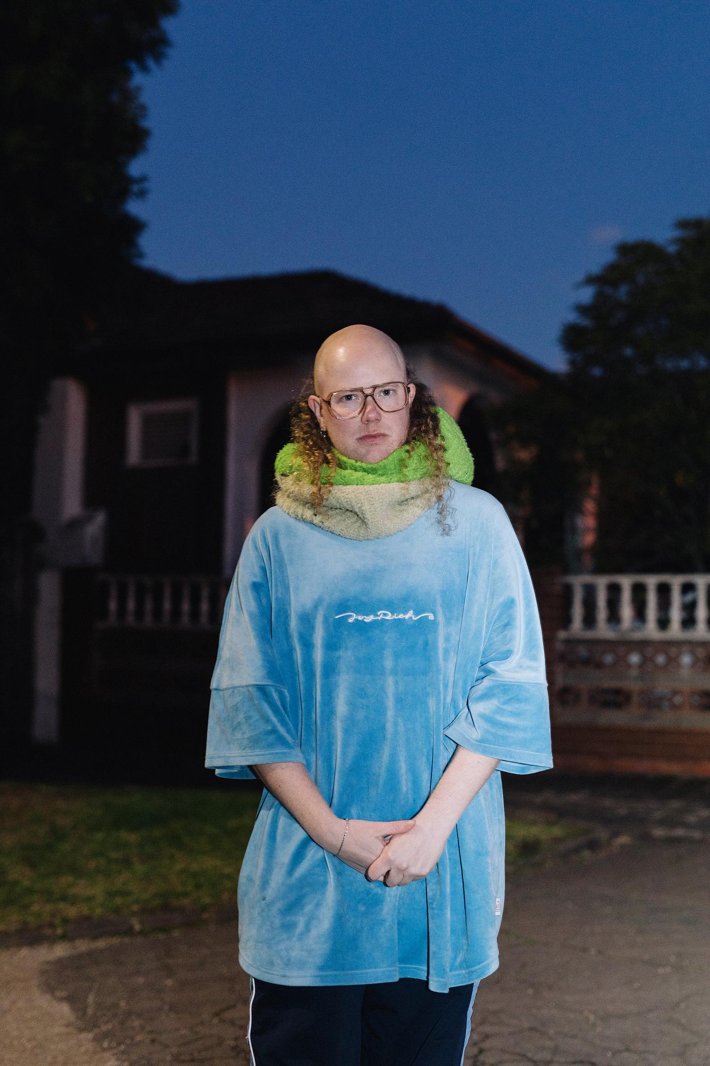 Archie Barry stands outdoors in front of a house at dusk, wearing a loose blue velour shirt and a large green scarf, with hands clasped in front.