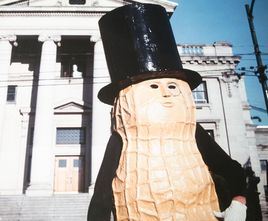A person wearing a peanut costume adorned with a face and a top hat stands in front of a civic building.