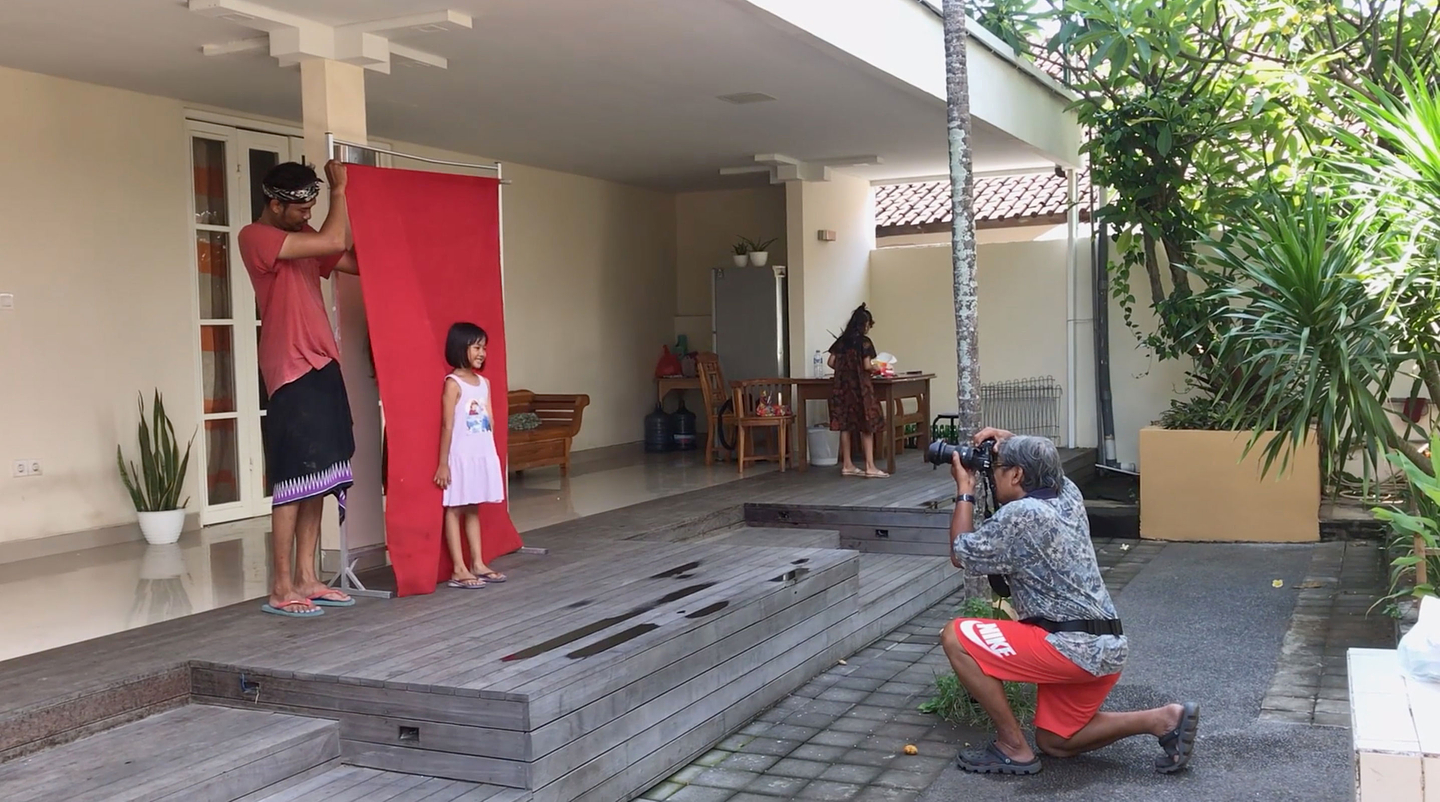 A man holds a red cloth backdrop up behind a small girl as they stand on a deck in a backyard. Another man crouches a few meters away from her, taking her picture as she faces the camera.