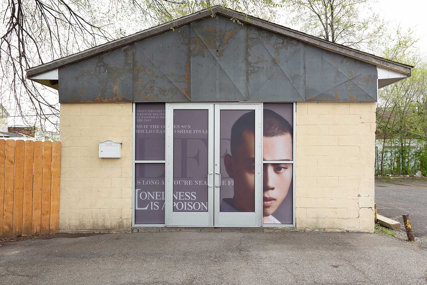 A portrait of Bruno Zhu wraps behind a metal door on a yellow brick storefront. Mimicking a spread in a fashion magazine, he appears against a purple background with white serif text, the most legible line reading Loneliness is a Poison. His skin looks airbrushed or as if run through a beauty filter.