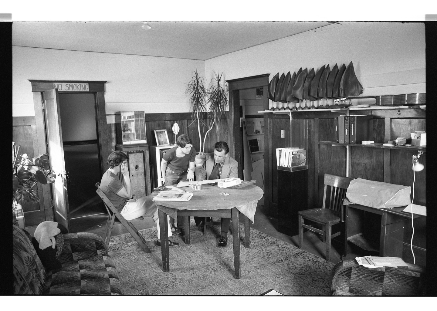 A black and white photo of Glenn Lewis, Kate Craig, and Jan Hoet gathering around a circular table in Western Front's library to look at documents. The library is furnished with upholstered couches, a patterned rug, artworks, and a number of shelves holding office supplies, binders, and a row of shark fin-shaped swimming caps.