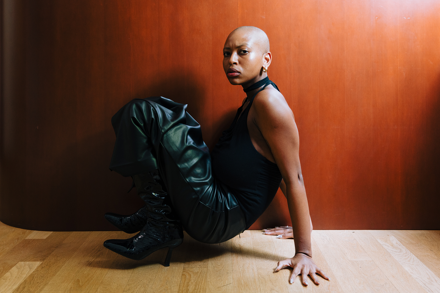 Facing parallel to the camera, Chipo Chipaziwa meets the camera’s gaze as she crouches on a wood floor. With her arms extended behind her, she balances her weight in her palms while her knees are bent towards her chest. She has a shaved head, and wears gold hoop earrings, a black halter top, leather skirt, and heeled patent boots.