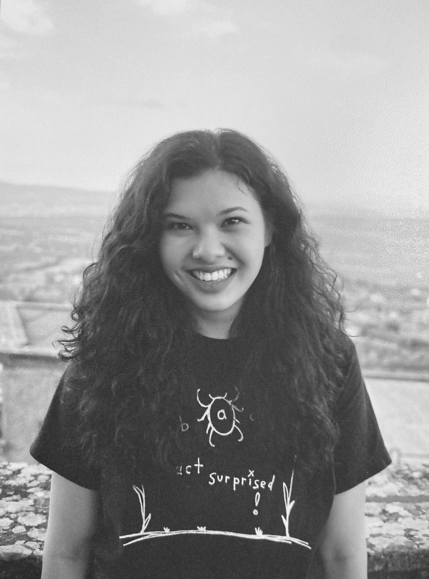 Black-and-white portrait of Kezia Elaschuk standing outdoors with a scenic, blurred landscape in the background. She has long, curly hair and is smiling warmly at the camera. She wears a graphic T-shirt with a whimsical insect drawing and the text "act surprised!" on it.


