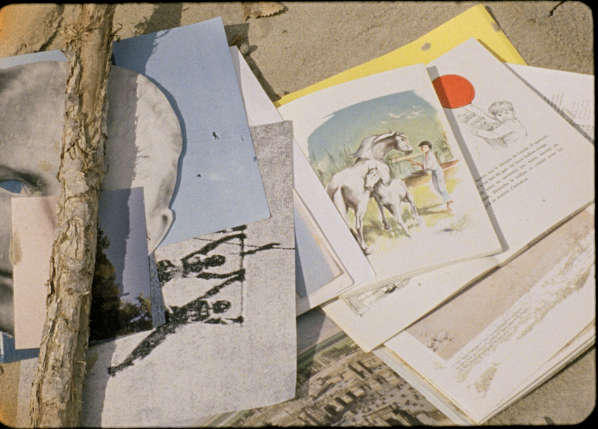 A collection of print ephemera scattered in the sand, including photographs, illustrations, and children's books, one of which shows a young boy with a red balloon. A tree branch lies across some of the papers. 