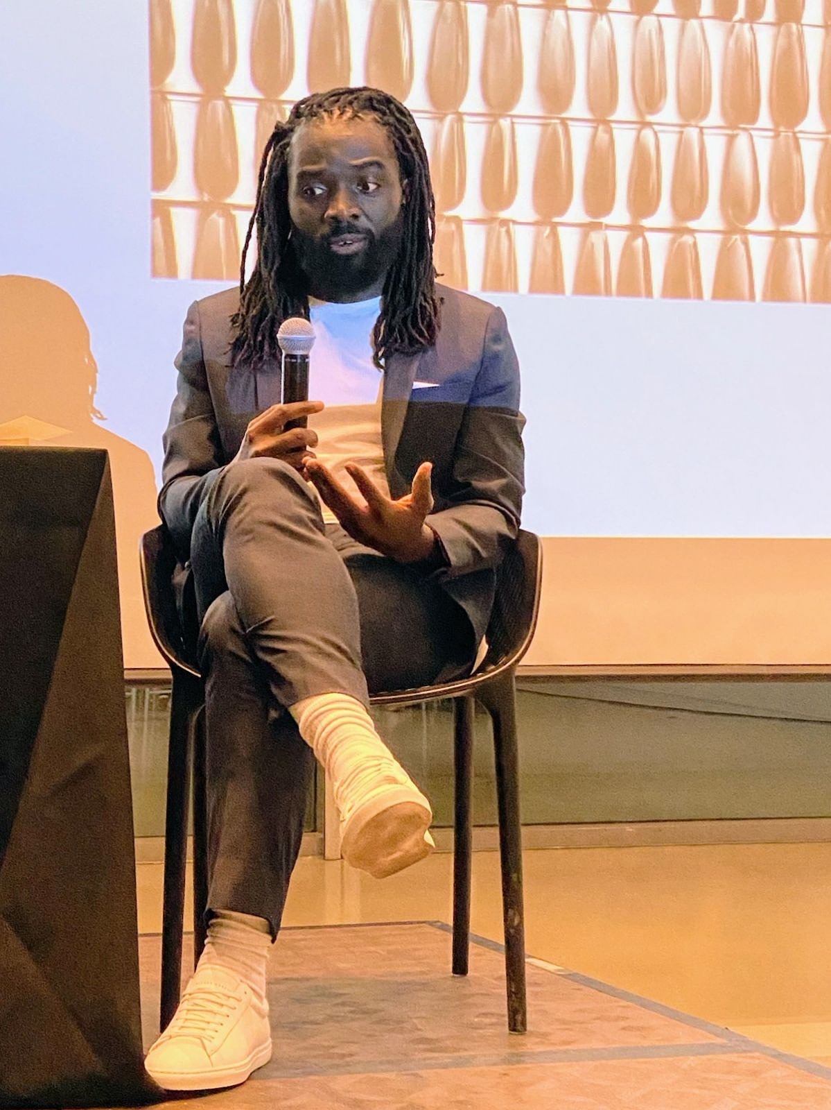 Phanuel Antwi sits on a black chair with one leg crossed over the other, speaking into a microphone. His expression appears thoughtful as he gestures with his free hand while addressing the audience. Behind him is a projected abstract pattern of repeated vertical shapes in warm tones. 