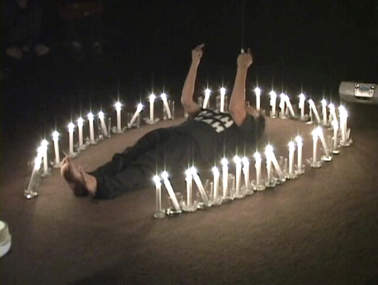 The artist Rebecca Belmore is dressed in black and lays on a carpeted floor with her arms in the air and her middle fingers raised. Surrounding her is an arch of lit candles. A silver boombox can be seen on the floor behind her.