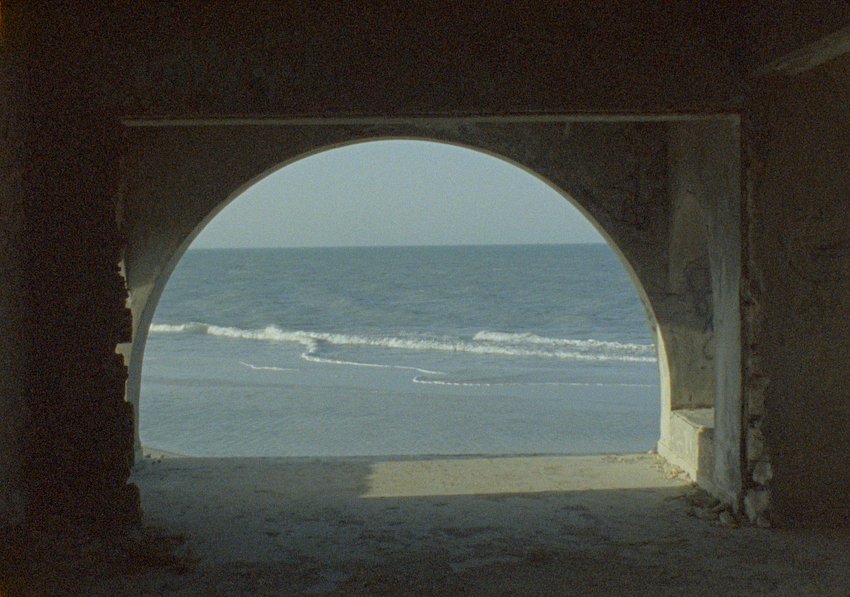 A peaceful coastal view seen through a large, arched opening in a weathered stone structure. The archway opens to the sea, with soft waves rippling onto the beach under a clear, light blue sky.