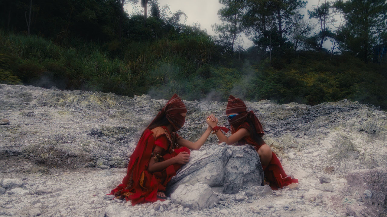 Two people dressed in red attire and face coverings are crouched on either side of a large rock. With their elbows balanced on the surface of the rock, they grip the others’ hand as if arm wrestling. Lush foliage can be seen beyond the rocky hill where they’re positioned. 
