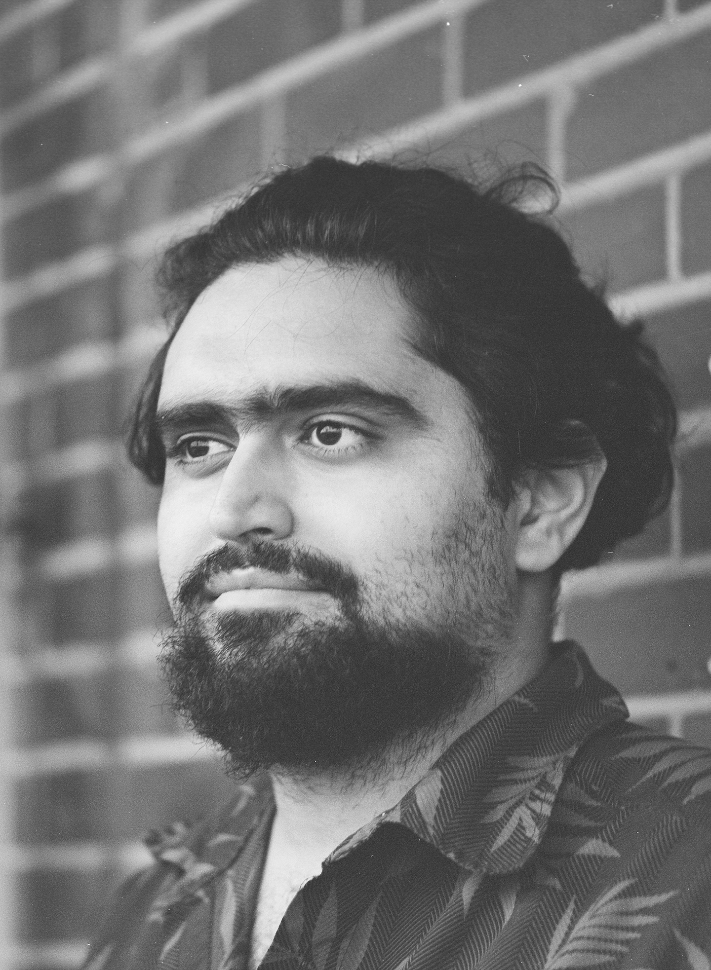 A black-and-white headshot of Khashayar Mohammadi at three-quarter view. They wear a collared shirt with a foliage pattern, and are positioned in front of a brick wall.