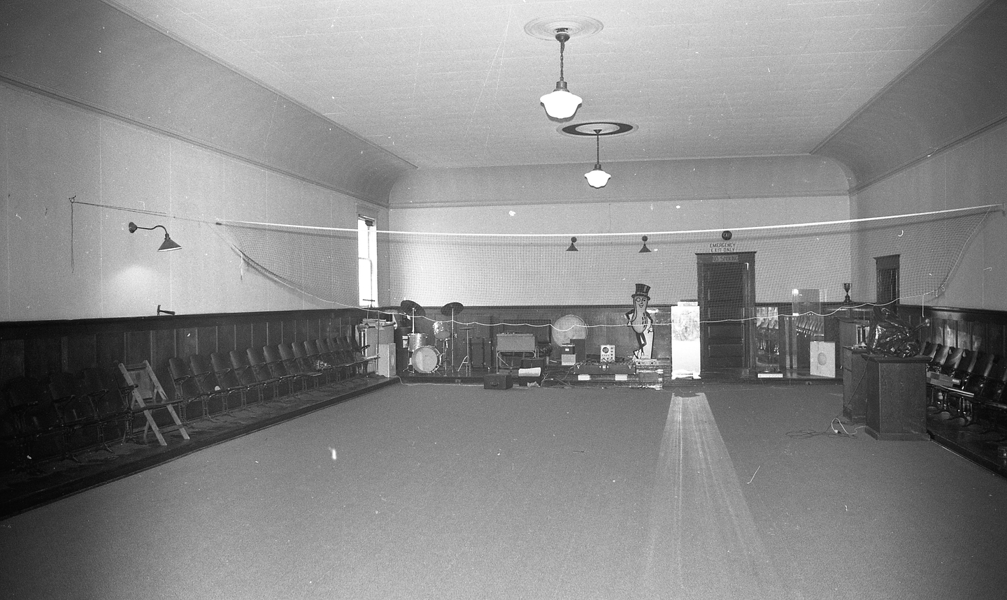 A black and white photo of the interior of Western Front’s Grand Luxe Hall, showing a view of the large room looking toward the back wall. The room is mostly empty apart from some equipment, including a drum set, lecterns, and large cardboard cut-out of Mr. Peanut.