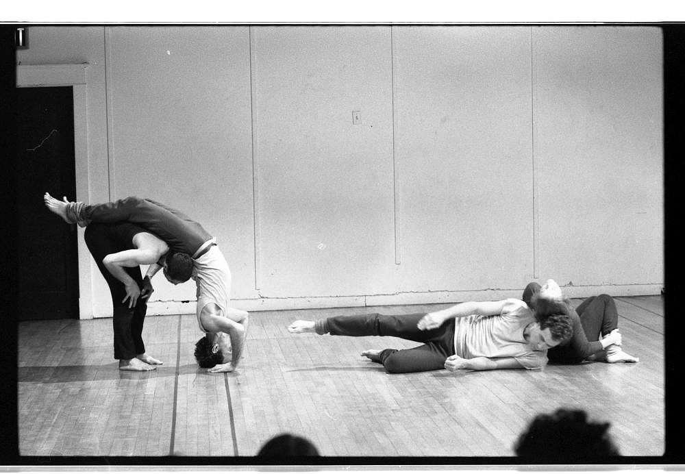 A black and white photograph of a dance performance. To the left Steve Paxton bends forward while Peter Bingham performs a headstand and arches his legs over Paxton's back. To the right Lola Ryan lays on her side and Helen Clarke leans against her back in a crouched position.