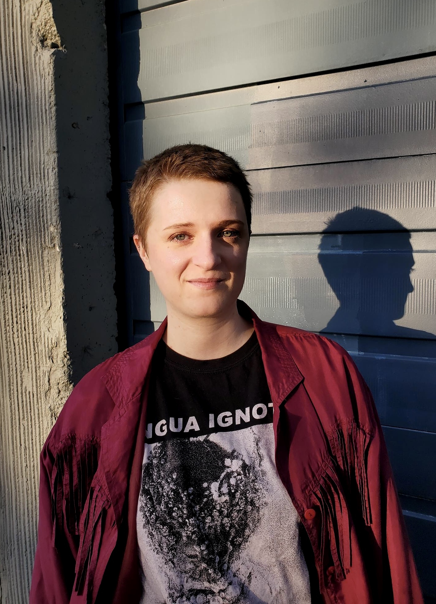 A portrait of Cristina Holman. She wears a black and white graphic t-shirt under a red button up shirt with tassels on the front and stands in front of a wooden vertical beam and green building siding.