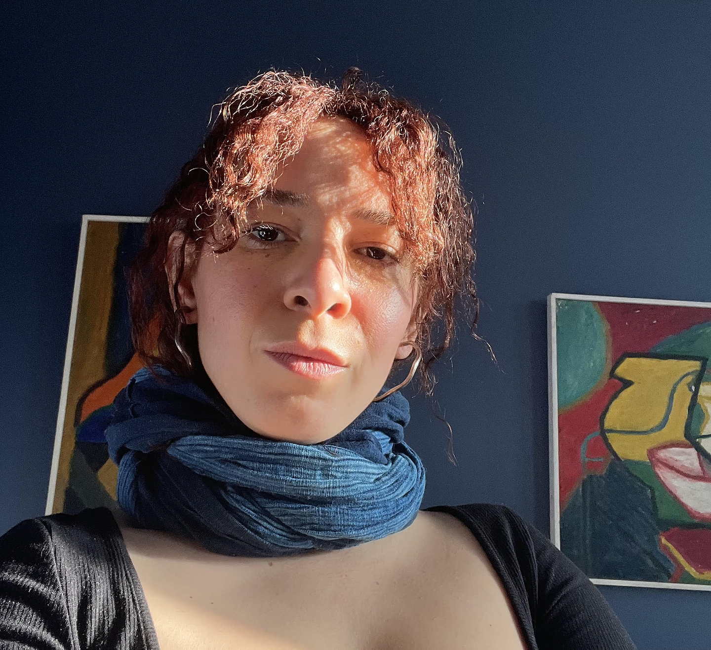 Zoe Imani Sharpe wears a blue scarf and black ribbed top and is lit by a ray of sun streaming in from somewhere out of frame. Behind her, two colourful abstract drawings hang on a blue wall.